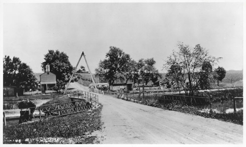 This photograph shows Washington Road in Princeton during an era of significantly reduced automobile traffic.  The building on the left is the bridgetender’s house, while the small structure on the right is the tender’s shelter.    The shelter provided the tender with a warm and dry place to await oncoming vessels.  At each bridge and lock along the Delaware and Raritan canal, the canal company built a home for the tender.  Although many of these buildings were demolished in the decades following the canal’s closure, a few of these historically-significant structures remain in places such as Lambertville, Trenton, Kingston, Lawrence, Griggstown, Blackwells Mills, Zarephath, and East Millstone.